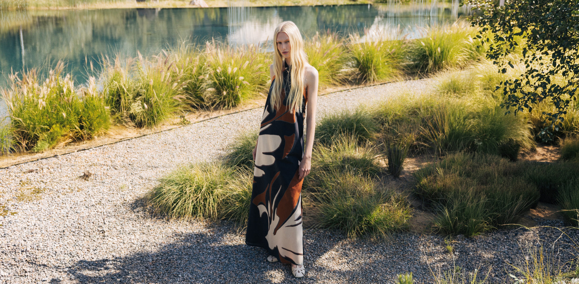 A woman stands on a gravel path outdoors near a lake, wearing a long sleeveless maxi dress with an abstract black, brown, and cream pattern.