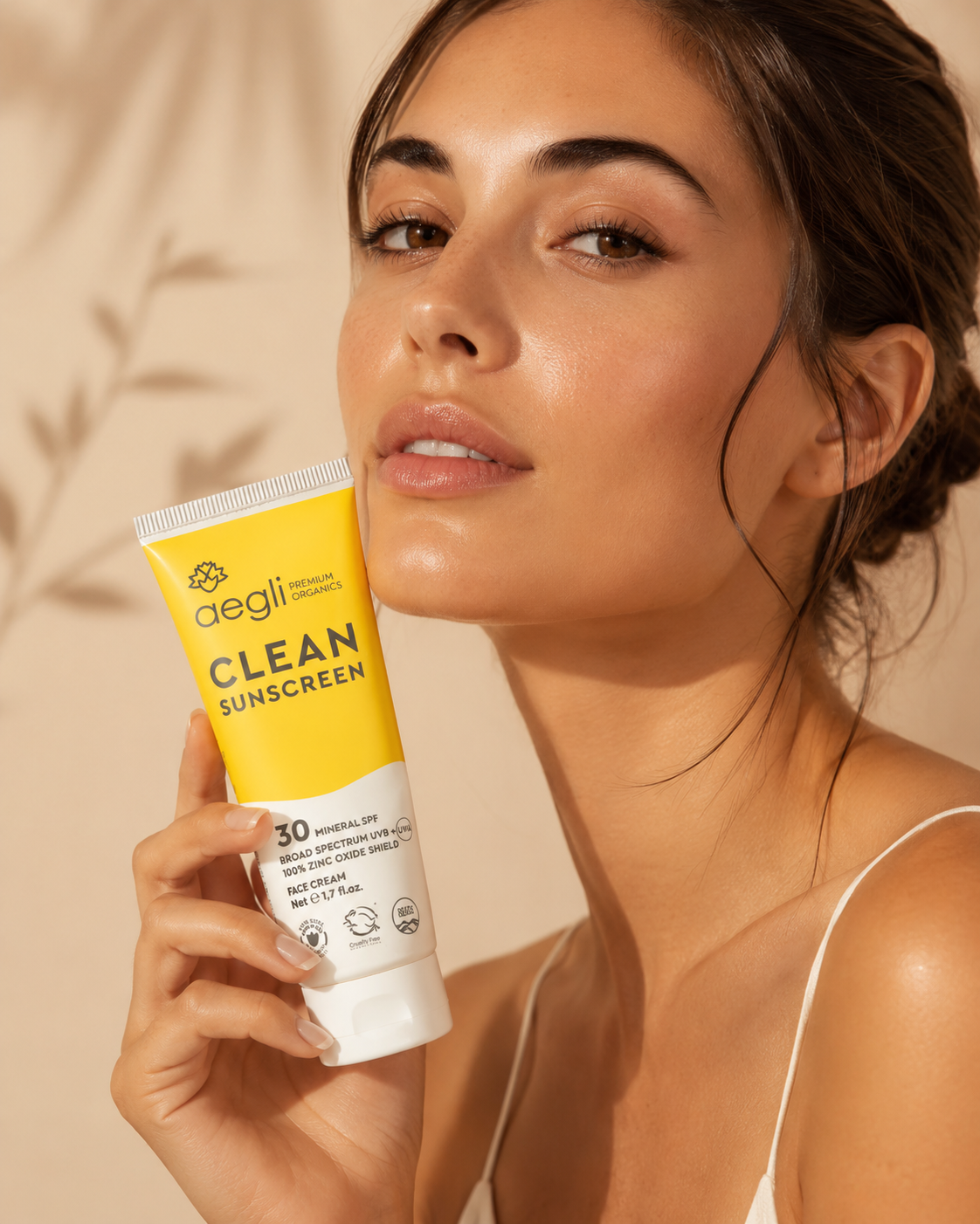 Model holding Aegli Clean Sunscreen SPF 30 in a yellow tube, shown in a close-up beauty shot with radiant skin and a neutral camisole.