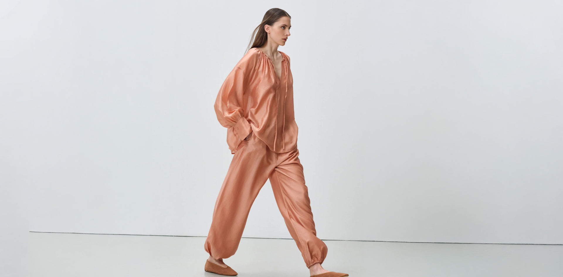 Model wearing a peach satin long-sleeve blouse with gathered cuffs and matching relaxed jogger pants with elastic ankles, styled with pointed flats.
