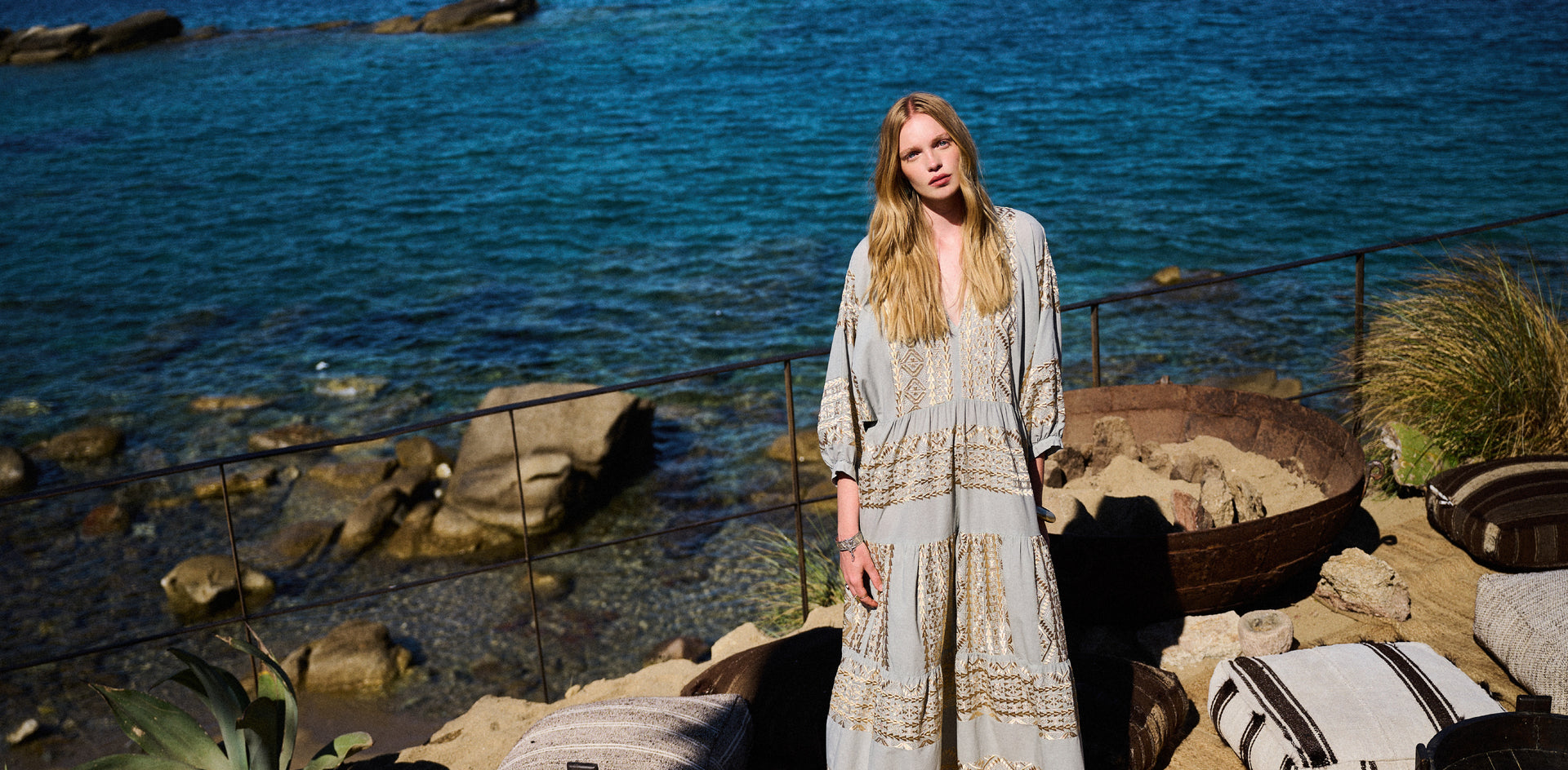 A woman stands on a rocky shoreline with blue water behind her, wearing a long light beige bohemian maxi dress with gold-toned embroidery. A striped pillow and other beach-style cushions are visible in the foreground.